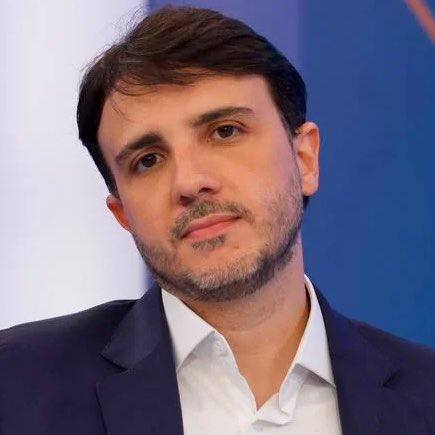 Homem falando, com expressão séria, em ambiente interno; aparece cercado por pessoas cuja postura sugere debate político.