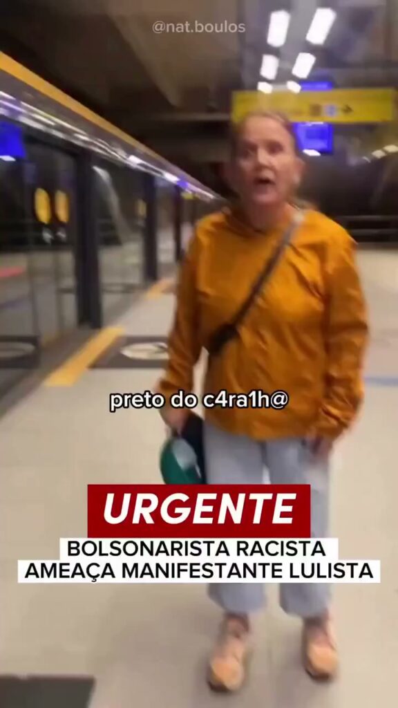 Pessoa em uma estação de metrô conversando com outras pessoas ao fundo; a cena parece acontecer perto de uma área de passagem com cores neutras.