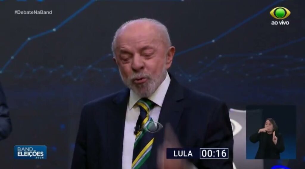 Foto de uma fala pública: homem em destaque, gesticulando com o dedo apontado, diante de microfones. Ao fundo, pessoas e ambiente de imprensa.